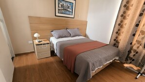 A bedroom with a wooden headboard, a single bed with a gray and red blanket, a nightstand with a lamp, a drawer, and a picture on the wall. - Cozy central shelter, steps away from the Mall (Ideal for families and friends). (Trujillo)