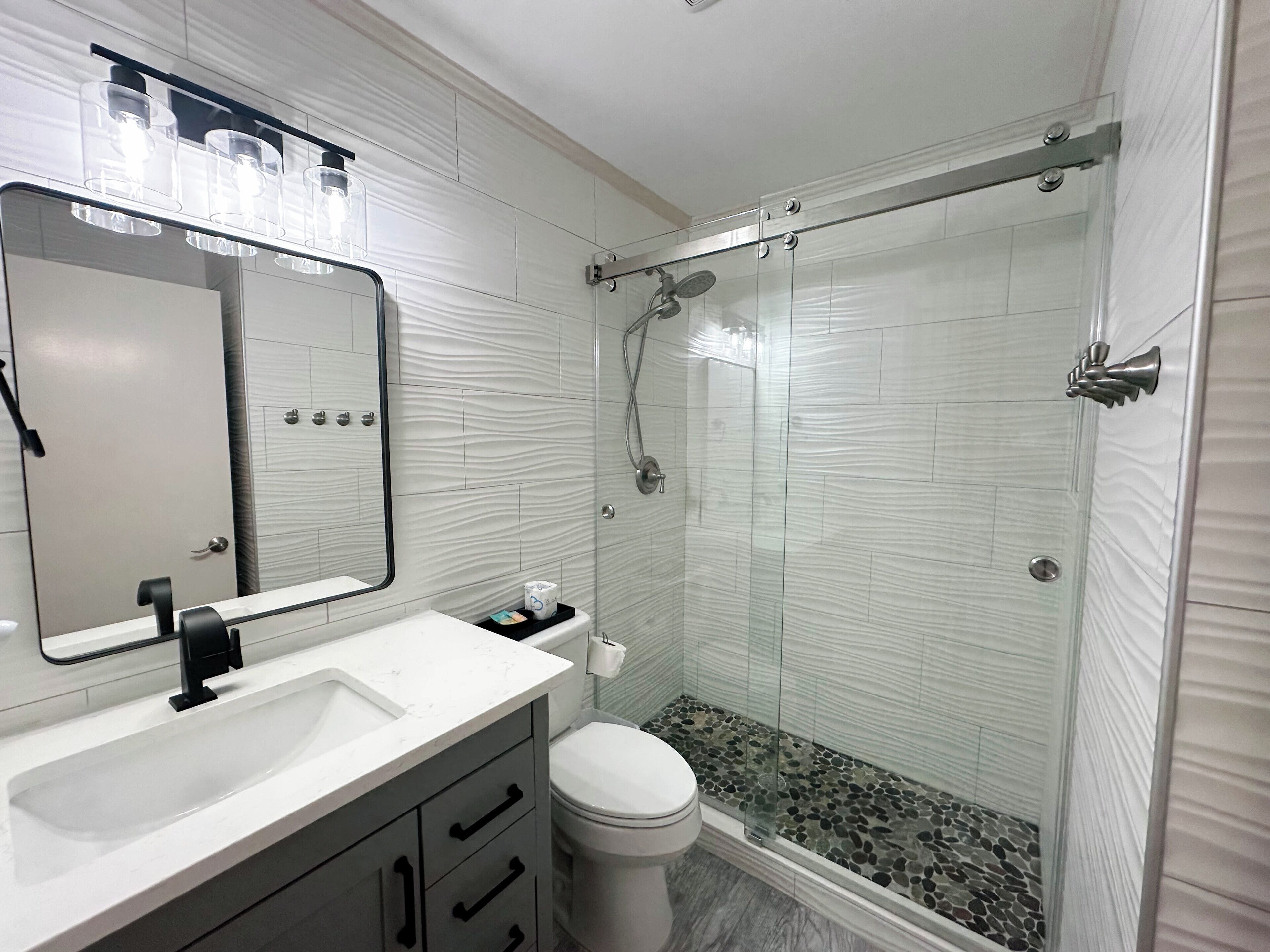 Un baño moderno con lavamanos, espejo y retrete blancos, ducha con mampara de vidrio y suelo de baño con piedras.