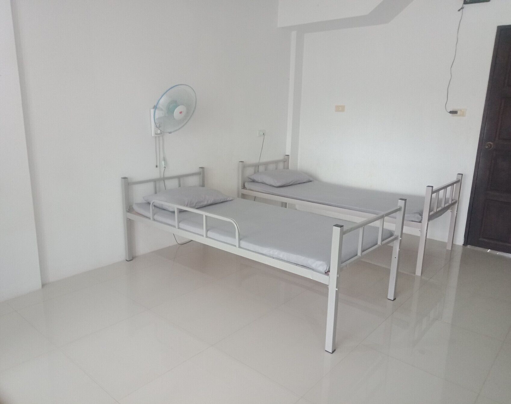 Une chambre avec un lit simple, un cadre en métal blanc, une petite table de chevet, un ventilateur fixé au mur et une porte avec un cadre foncé.
