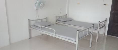 Une chambre avec un lit simple, un cadre en métal blanc, une petite table de chevet, un ventilateur fixé au mur et une porte avec un cadre foncé.