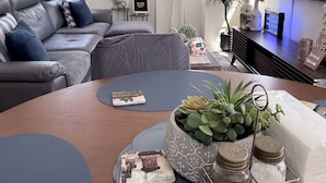 A modern living room with a grey sofa, a round coffee table with decorative items, a flat-screen TV, and a large window with sheer curtains. - Balaycation 3 BR, 2BA, 5 Beds, sleeps 8 (Davao City)