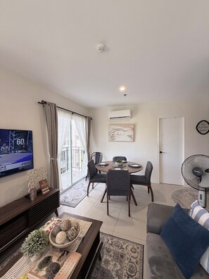 Een woonkamer met een televisie, een eettafel, stoelen, een bank, een ventilator en een deur.