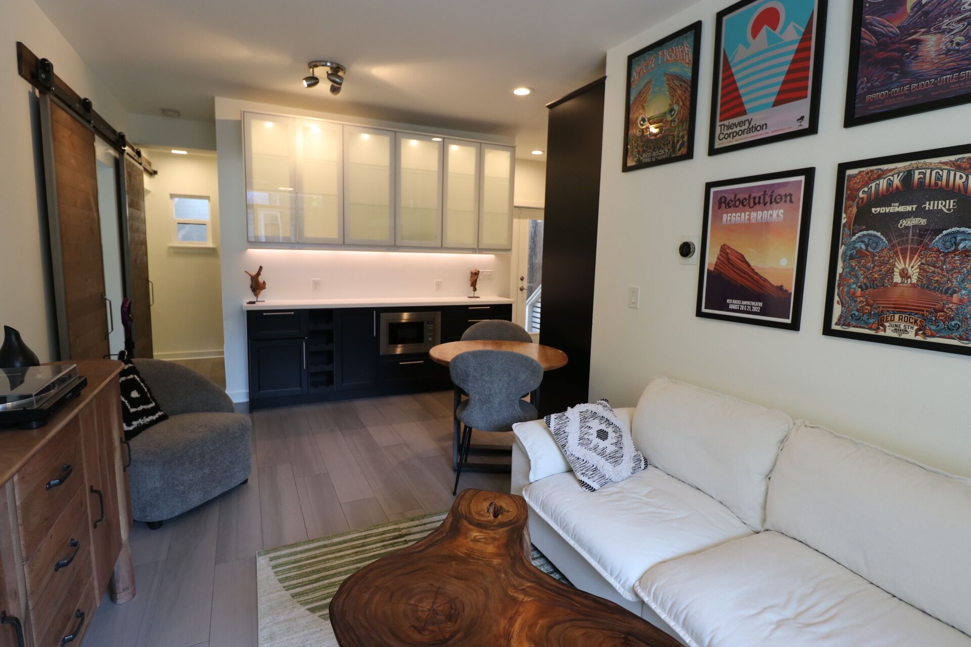 A modern living room with a white sofa, a wooden coffee table, and a grey armchair. A kitchen with white cabinets and a microwave is visible in the background.