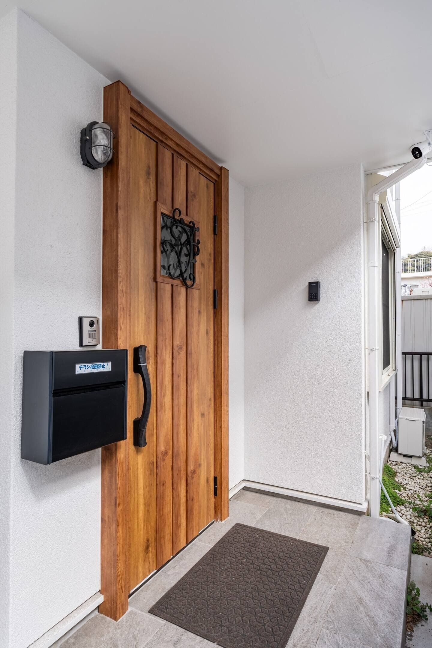 Een houten deur met een keypad-slot, een intercom bevestigd aan de muur, en een klein buitenruimte zichtbaar door een geopende deur.
