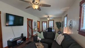 A living room with a large flat-screen TV, a ceiling fan, a dark-colored sectional sofa, and a wooden coffee table. - Vicky's Place - 3 bed, 1-bath, Pet Friendly w/Yard & Parking (Great Falls)