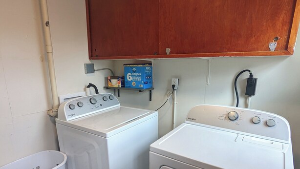 A laundry room with two front-loading washers and dryers, a wooden cabinet above, a white laundry basket, and a wall-mounted electrical outlet. - Vicky's Place - 3 bed, 1-bath, Pet Friendly w/Yard & Parking (Great Falls)