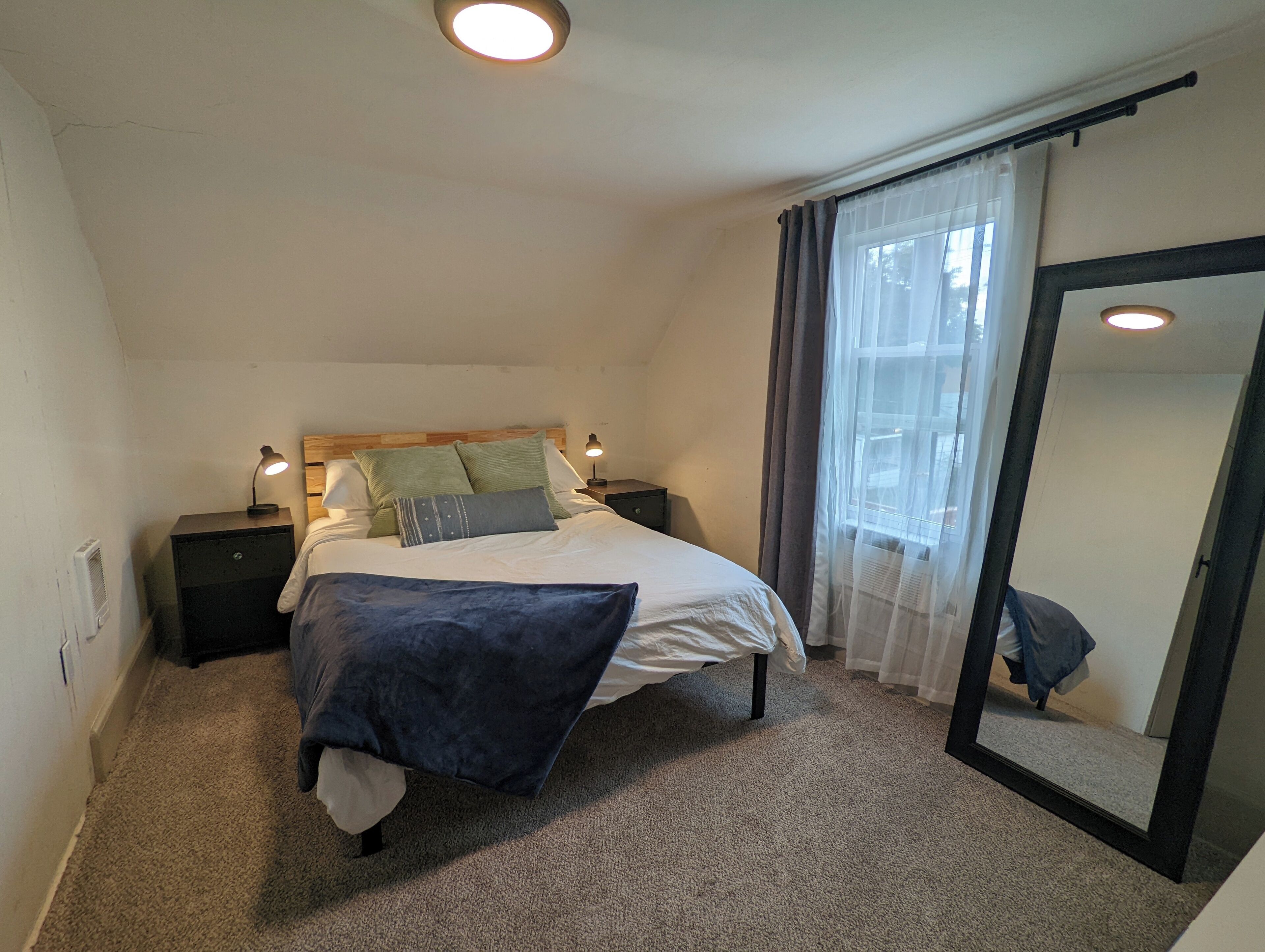 Une chambre à coucher avec un lit, des tables de chevet, un miroir et une fenêtre avec des rideaux.