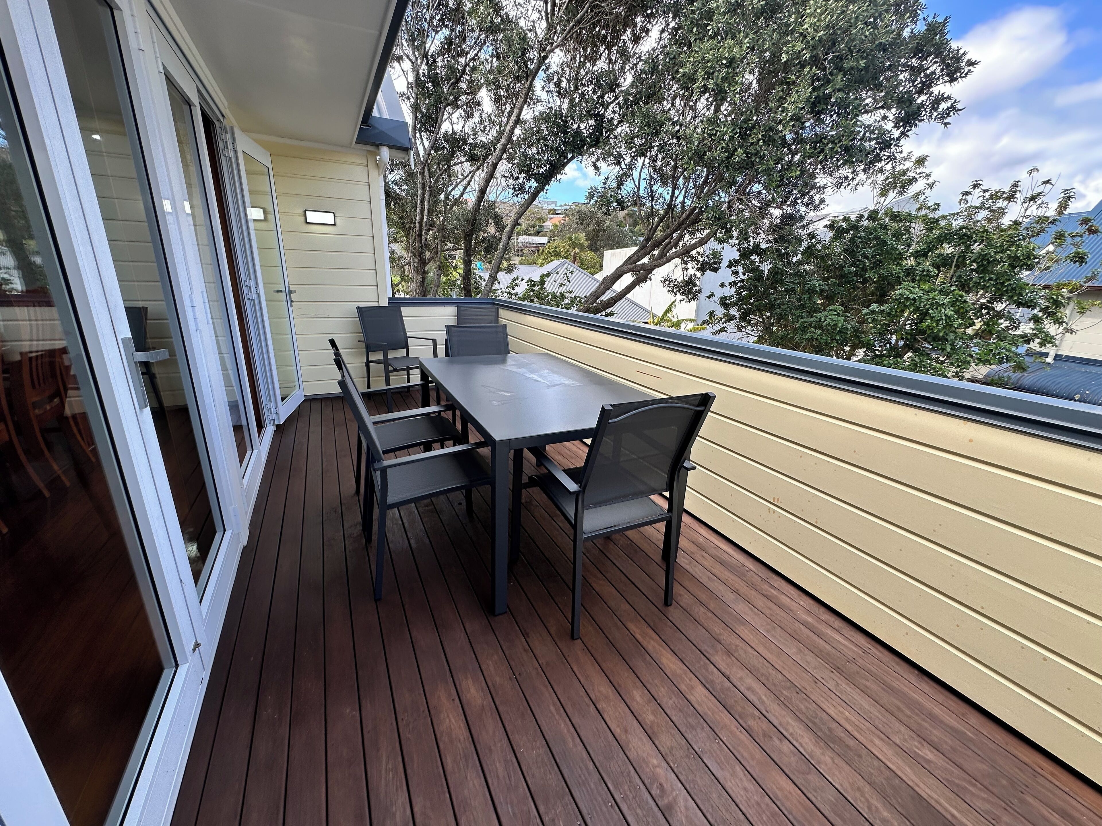 Una terraza de madera con una mesa y sillas, una puerta corrediza de vidrio y vistas a árboles y edificios.