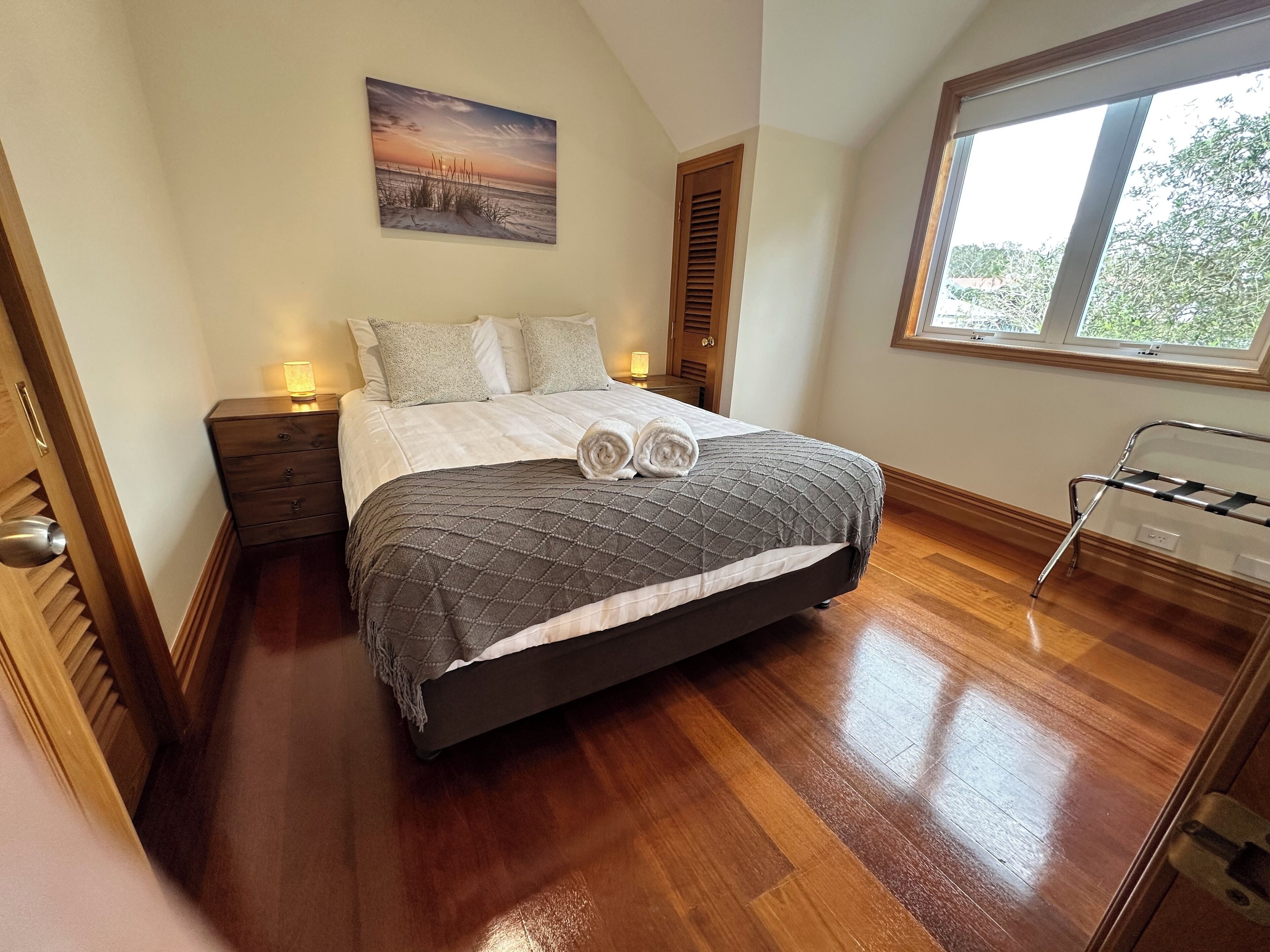 Een slaapkamer met een bed, nachtkastje, raam en een houten vloer.
