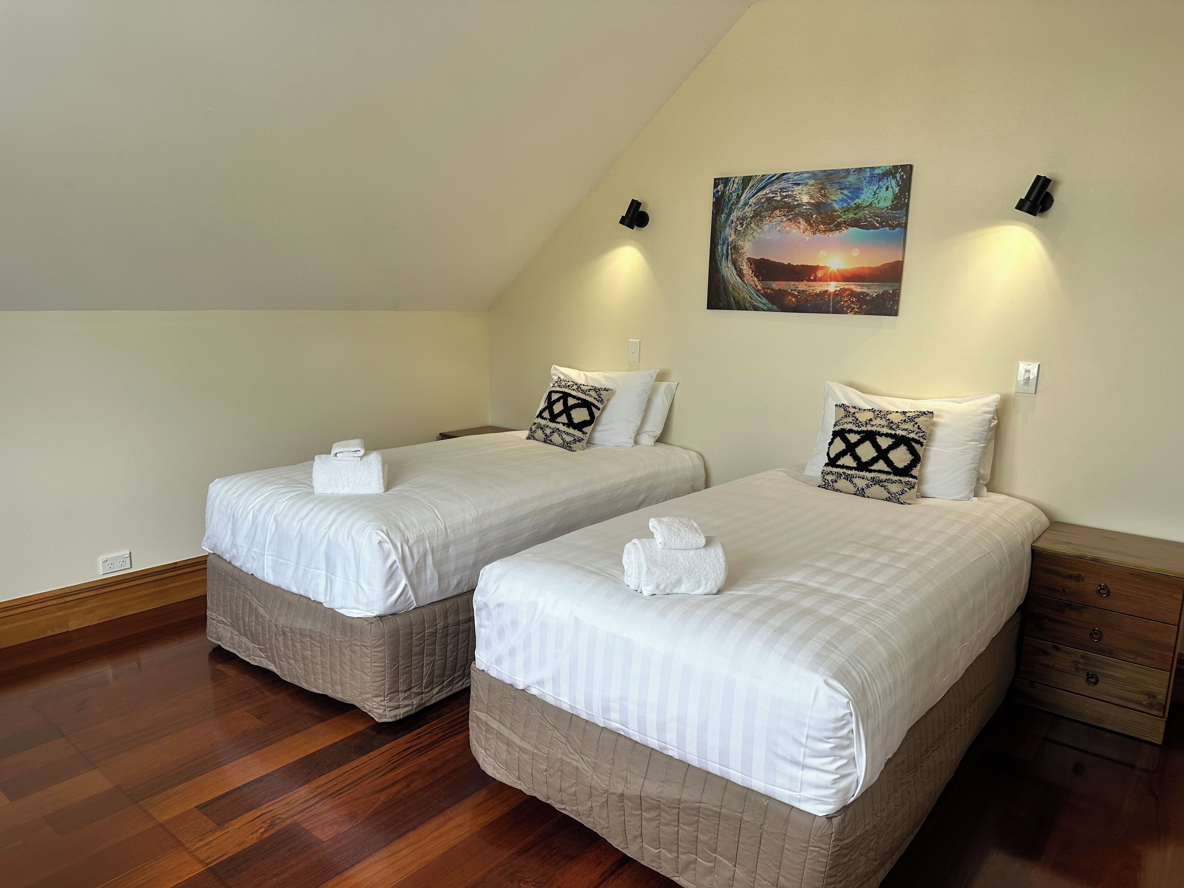 A room with two beds, each with a white bedspread and a towel folded on top. There is a painting of a sunset on the wall above the beds.