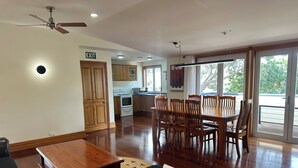 A spacious living area with a wooden dining table and chairs, a sofa, and a coffee table. There is a kitchen in the background with wooden cabinets and a microwave.