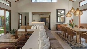 A spacious room with a long dining table, a sofa, and a kitchen area visible through an open doorway. - Villa Emmy | Elegant Seminyak 4BR Villa — Pool & Private Chef (Seminyak)