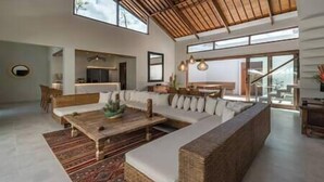 A spacious living area with a large sofa, a coffee table, and a dining table. There is a kitchen area in the background, and the room has large windows allowing natural light. - Villa Emmy | Elegant Seminyak 4BR Villa — Pool & Private Chef (Seminyak)