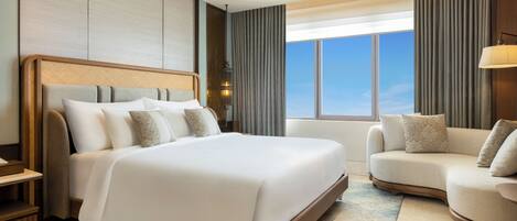Habitación de hotel con una cama grande, un sofá, una mesita de noche, una lámpara y vistas al cielo.