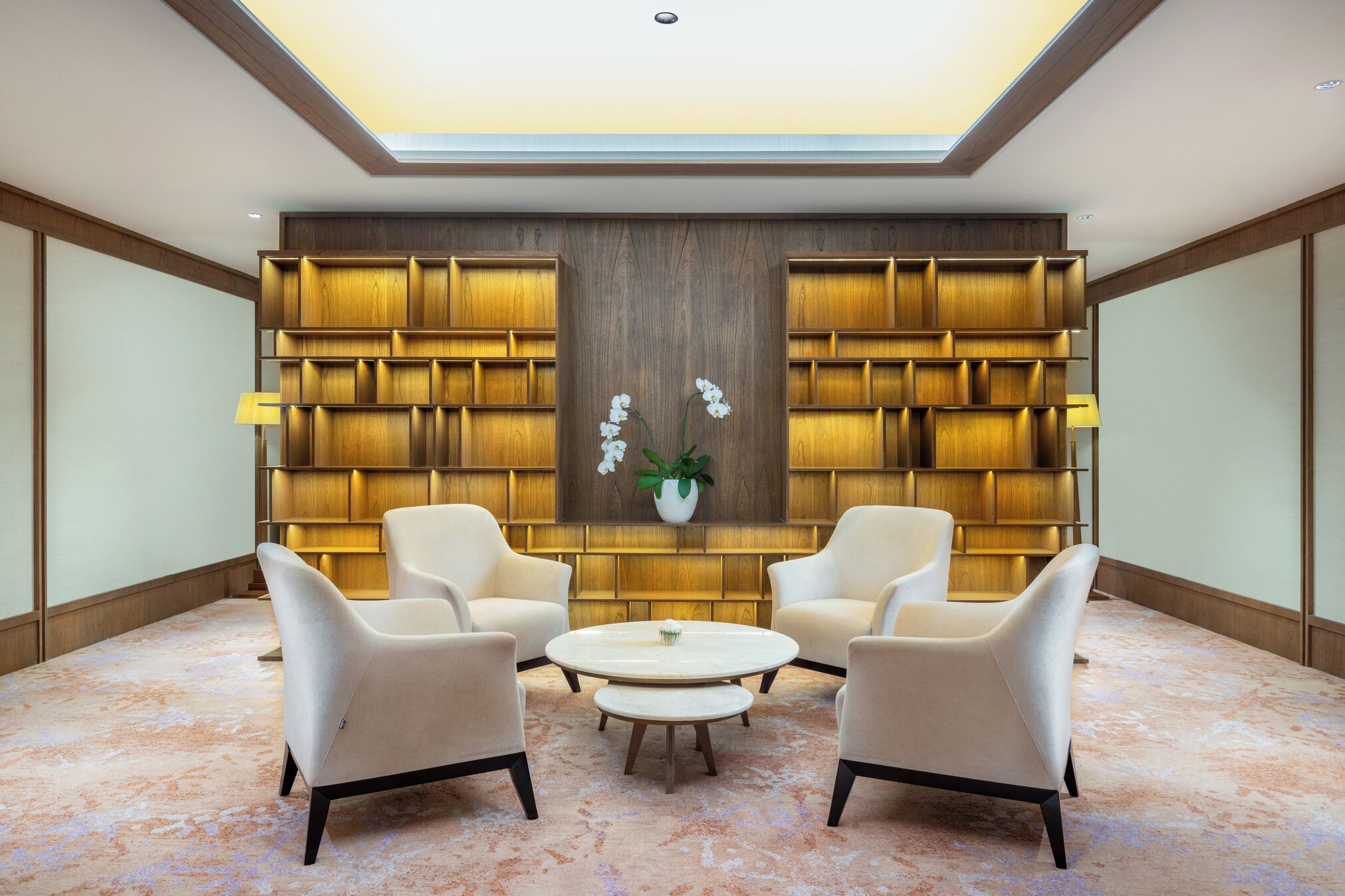 A modern lobby with a wooden bookshelf, two armchairs, a round coffee table, and a potted orchid.