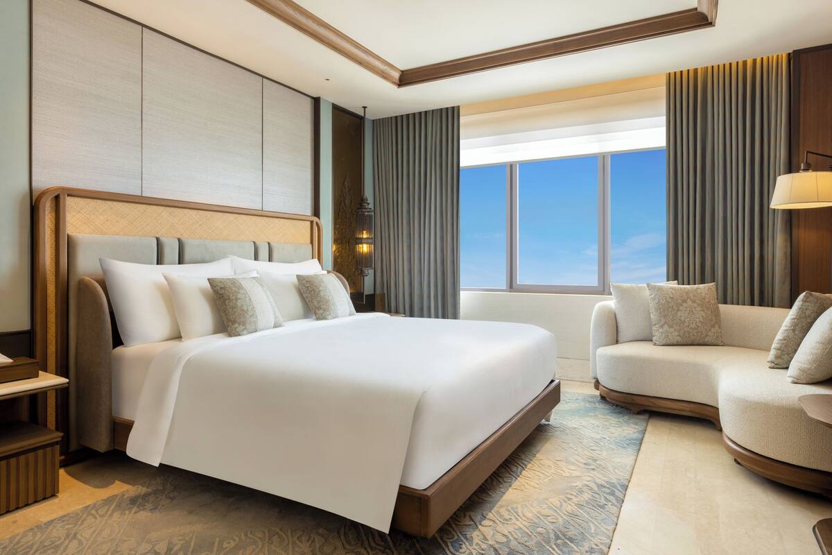 A hotel room with a large bed, a sofa, a nightstand, and a view of the sky.