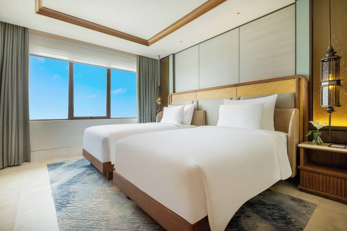 A hotel room with two beds, a large window, a nightstand with a lamp, and a decorative wall panel.