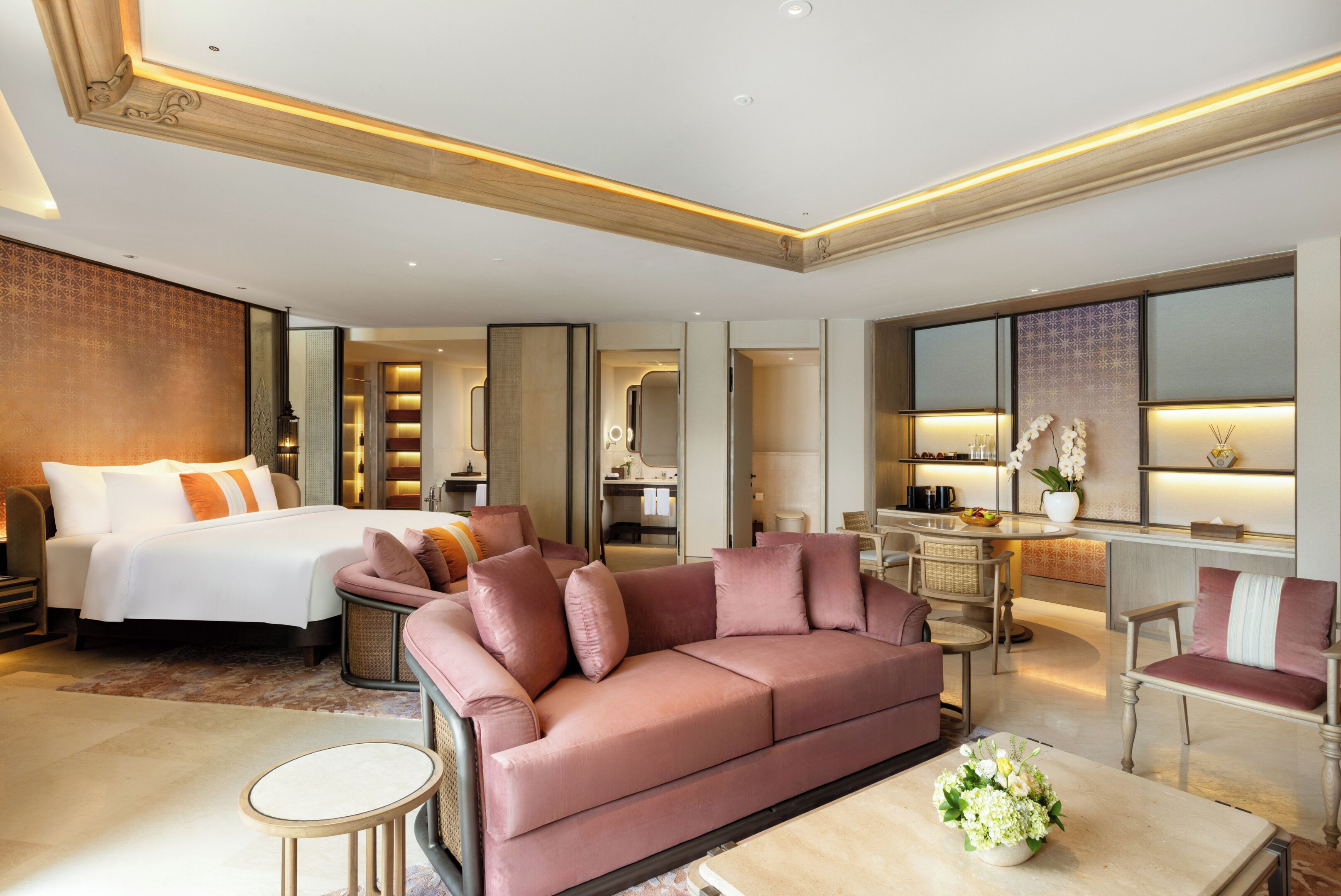A modern hotel room with a large bed, a pink sofa, a dining area, and a small table with flowers.
