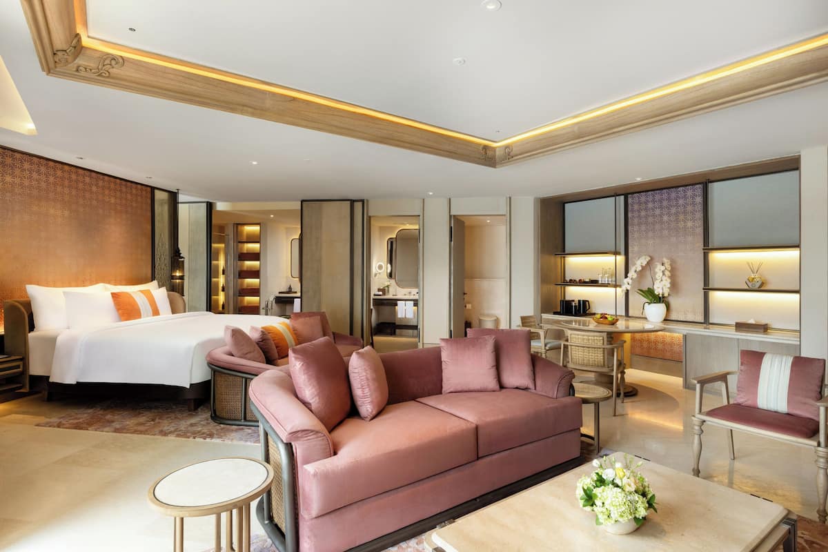 A modern hotel room with a large bed, a pink sofa, a dining area, and a workspace.