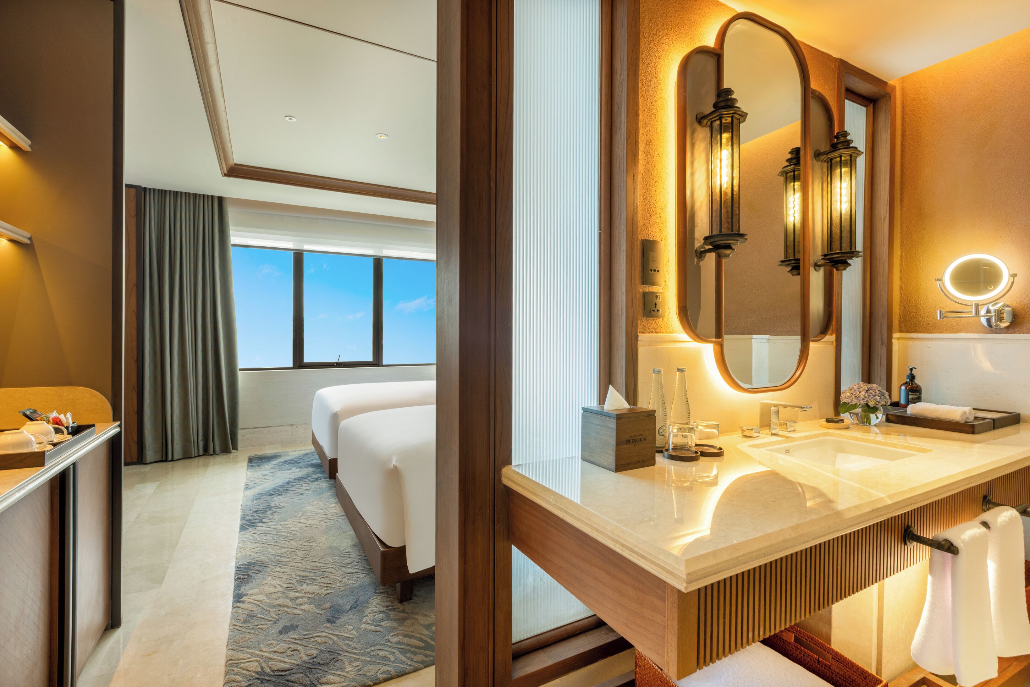 A hotel room with a large bed, a bathroom with a double sink and a large mirror, and a view of the sky through the window.