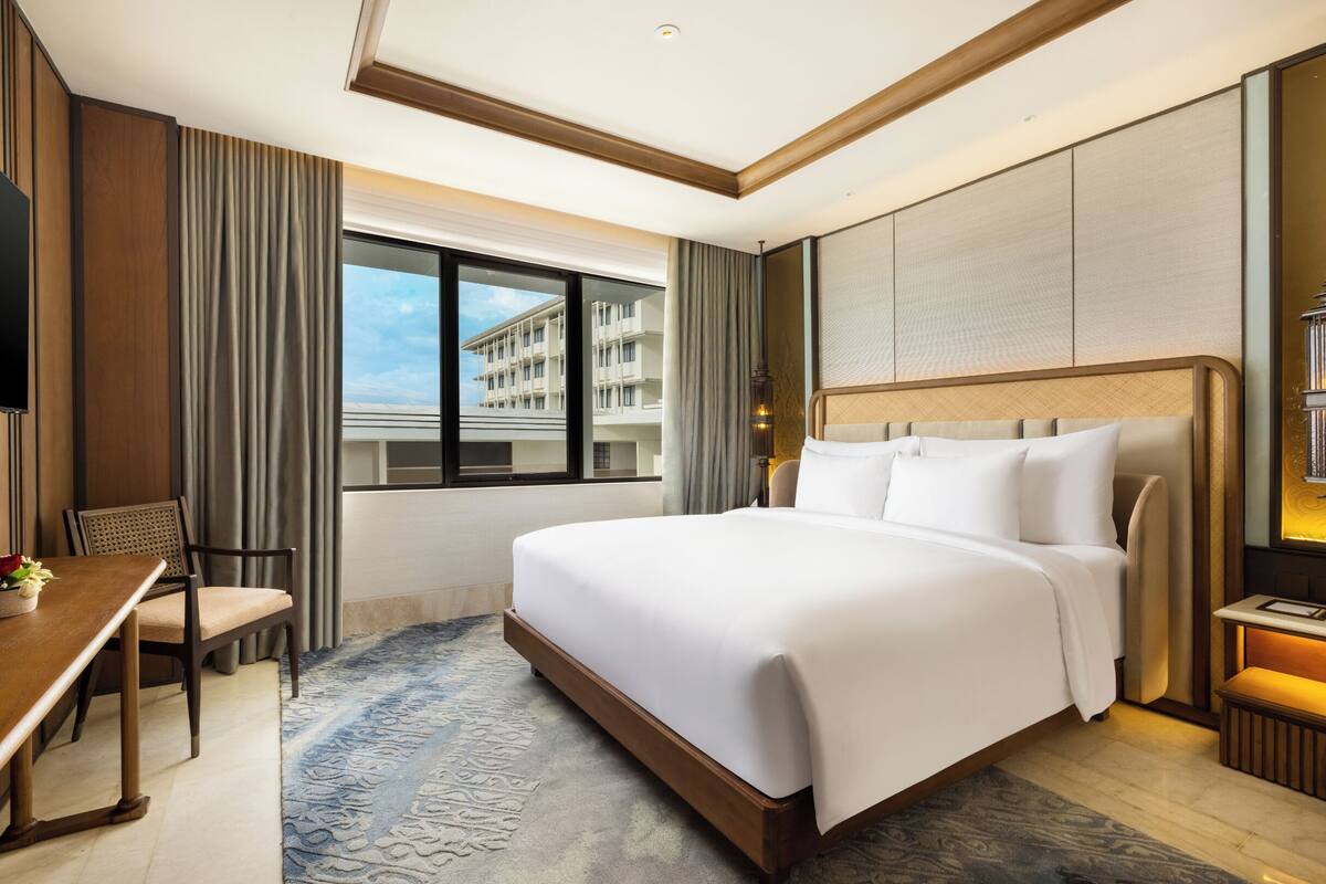 A hotel room with a large bed, a desk, a chair, a window with a view of buildings, and a small table with a lamp.