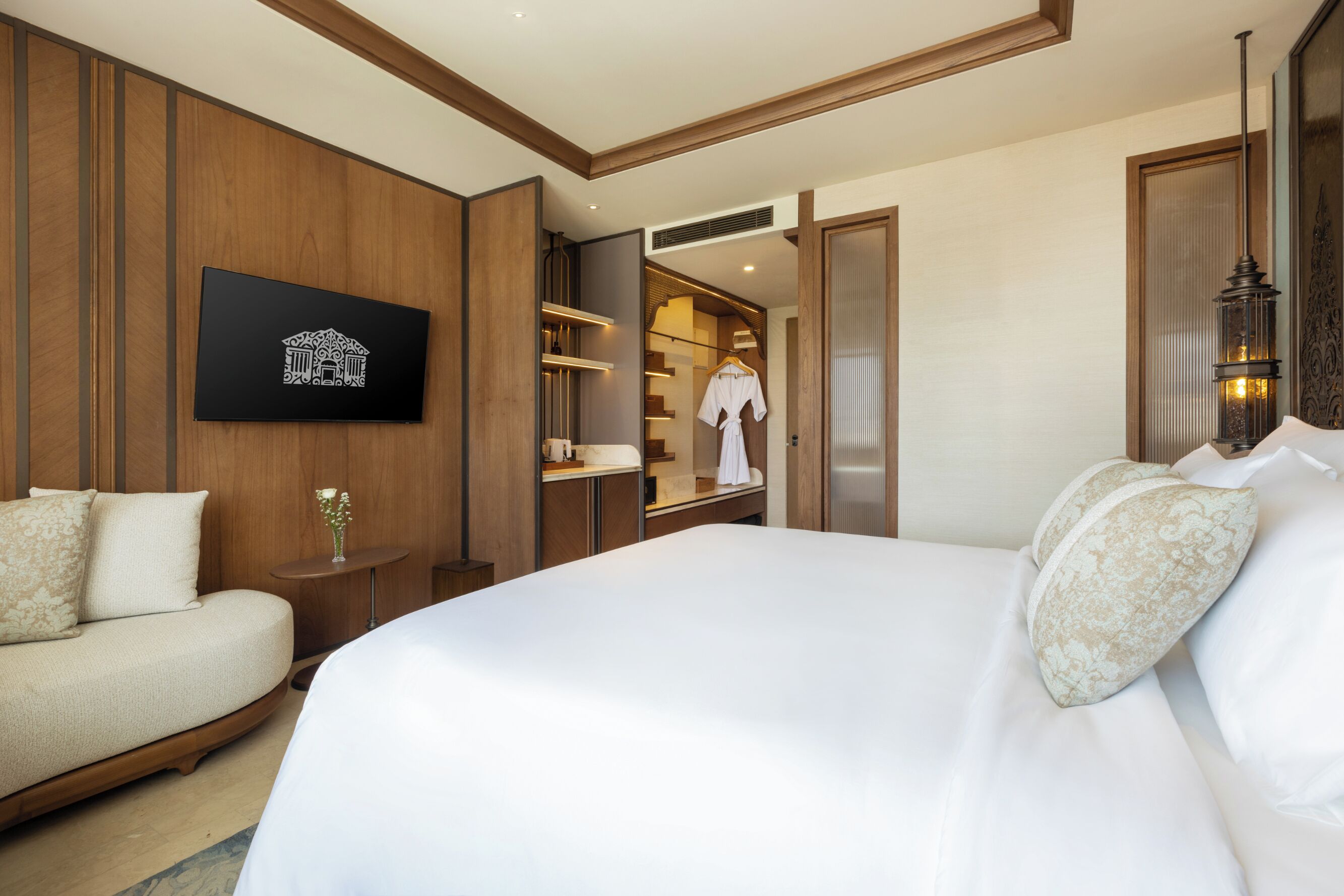 A hotel room with a large bed, a sofa, a small table with a vase, and a wardrobe with a robe hanging.