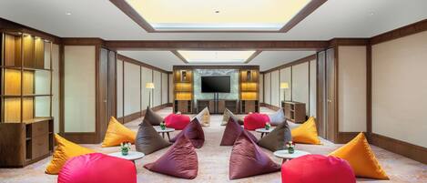 A spacious room with a large flat-screen TV mounted on the wall, multiple bean bags in various colors, and a high ceiling with skylight.