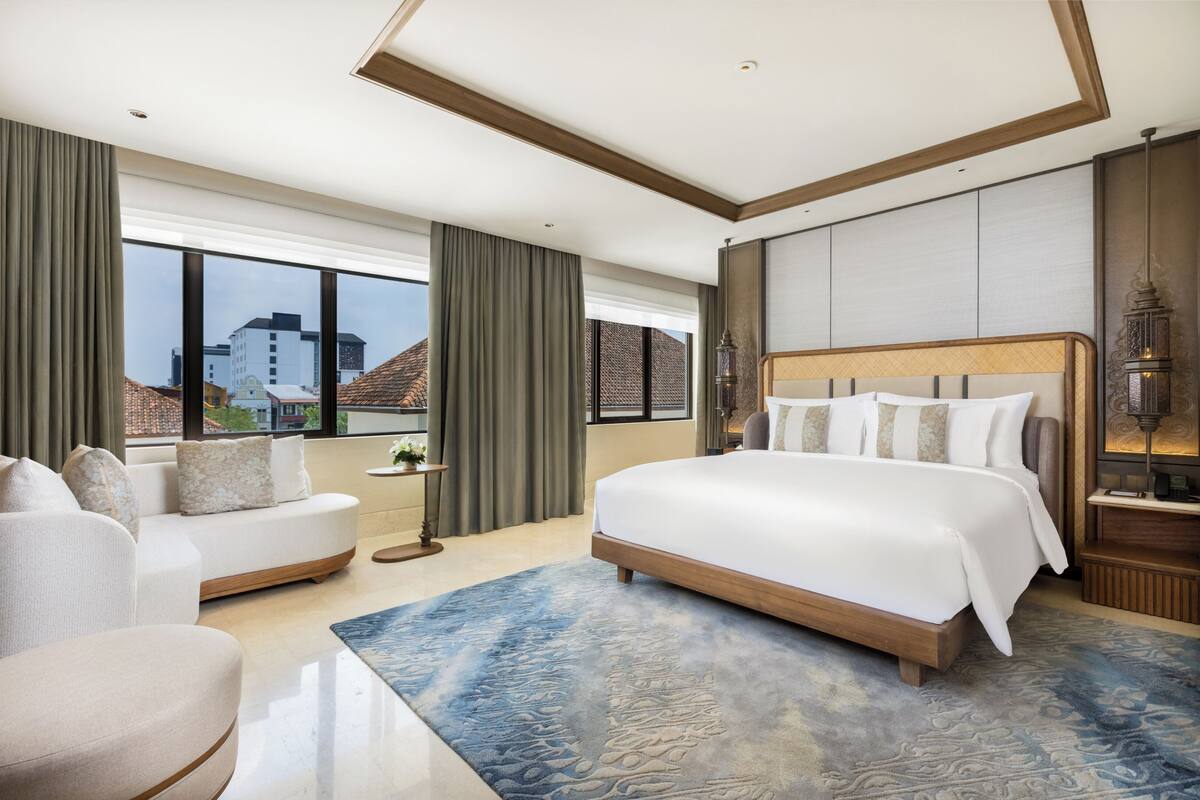 A modern hotel room with a large bed, a sofa, a small table with a plant, and a view of buildings through large windows.