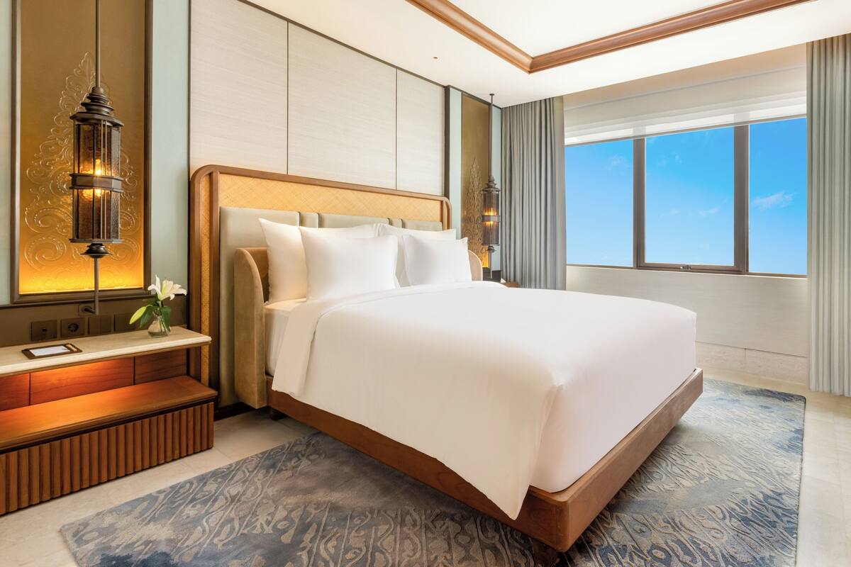 A hotel room with a large bed, bedside tables, a nightstand, a lamp, a view of the sky, and a window.