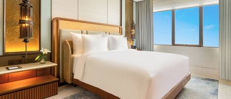 A hotel room with a large bed, bedside tables, a nightstand, a lamp, a view of the sky, and a window.