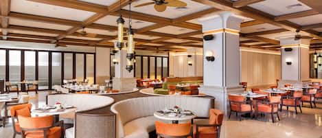Un comedor de hotel espacioso con diversas disposiciones de asientos, incluyendo mesas redondas y un sofá curvo, bajo un techo alto con vigas de madera y ventiladores.