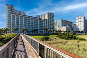 Un hotel con varios edificios altos, un paseo marítimo de madera y una zona verde con palmeras.