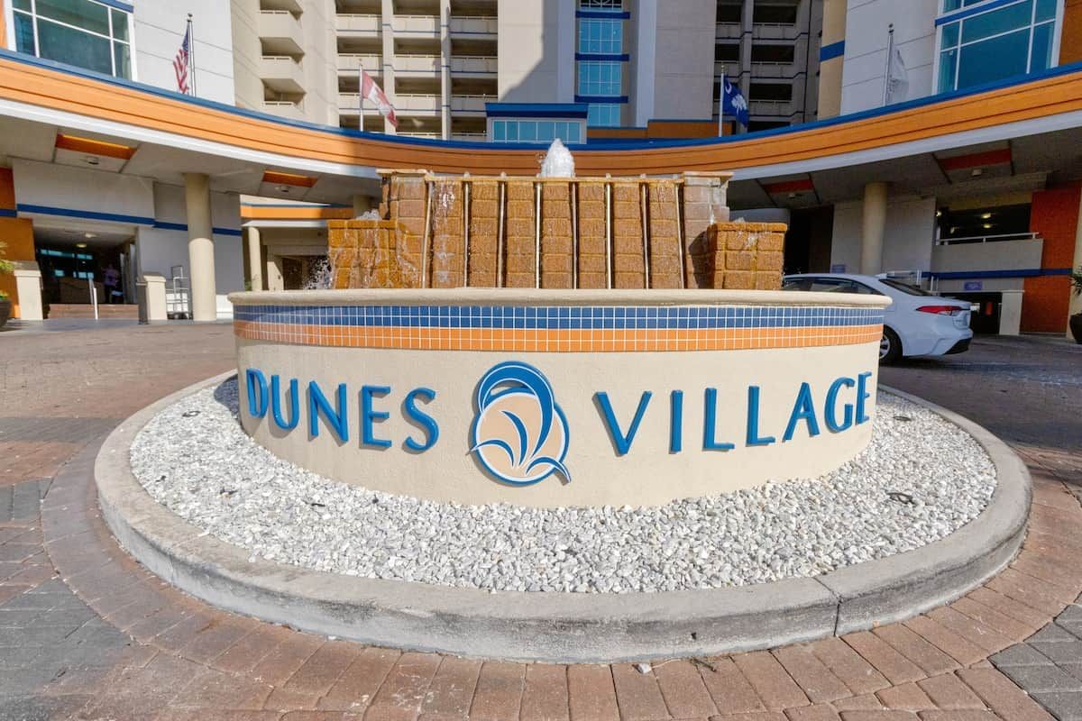 Sinal circular com o texto 'DUNES VILLAGE' e um logotipo, rodeado por cascalho e uma área pavimentada.