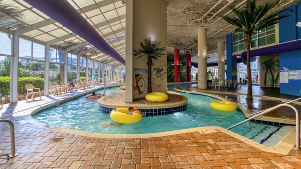 Une piscine intérieure dotée de plusieurs couloirs de nage, d’un îlot central avec un palmier, et de chaises longues disposées tout autour.