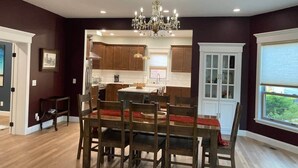 A dining area with a chandelier, wooden dining table, and chairs. There is a kitchen in the background with wooden cabinets and a window.