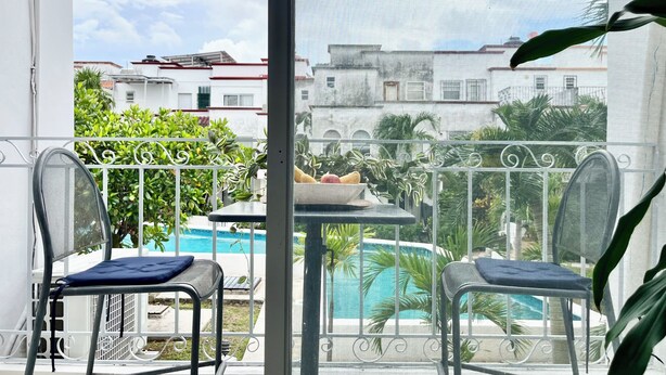 A balcony with two chairs, a small table with a bowl of fruit, and a view of a swimming pool and buildings. - Local atmosphere great location 2 bedroom, Fast WiFi Ideal for long stays. (Cancún)