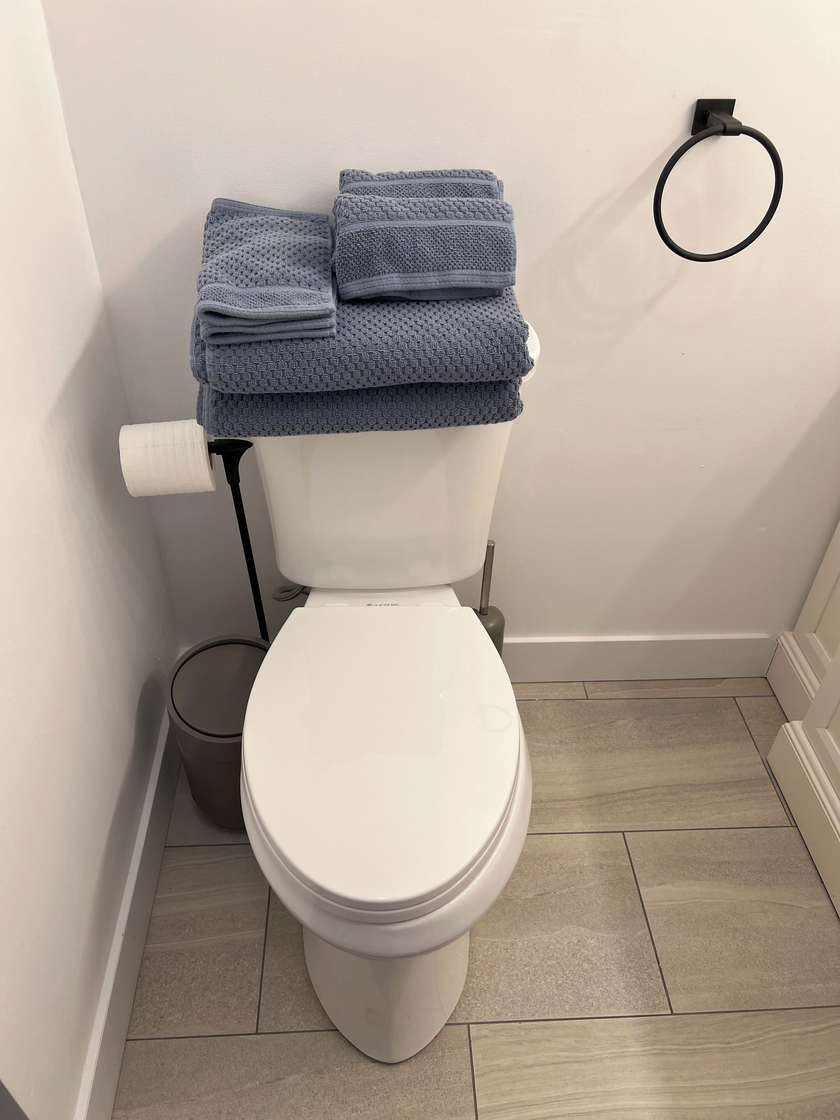 A toilet with a white lid, a roll of toilet paper, and a stack of folded towels on top.