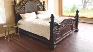 Una cama grande con cabecera y piecera de madera oscura, una mesita de noche de vidrio y vistas a un árbol a través de un ventanal amplio.