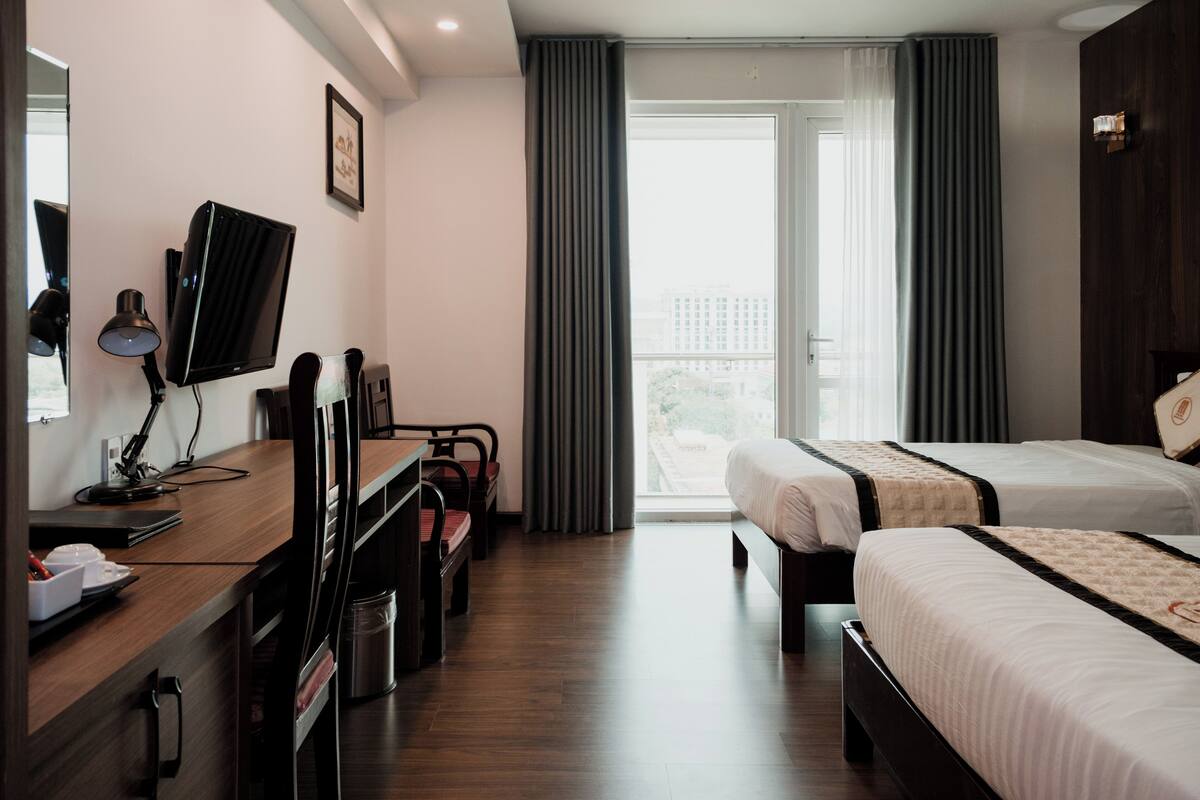 A hotel room with two beds, a desk with a computer, a chair, a TV, and a large window with curtains.