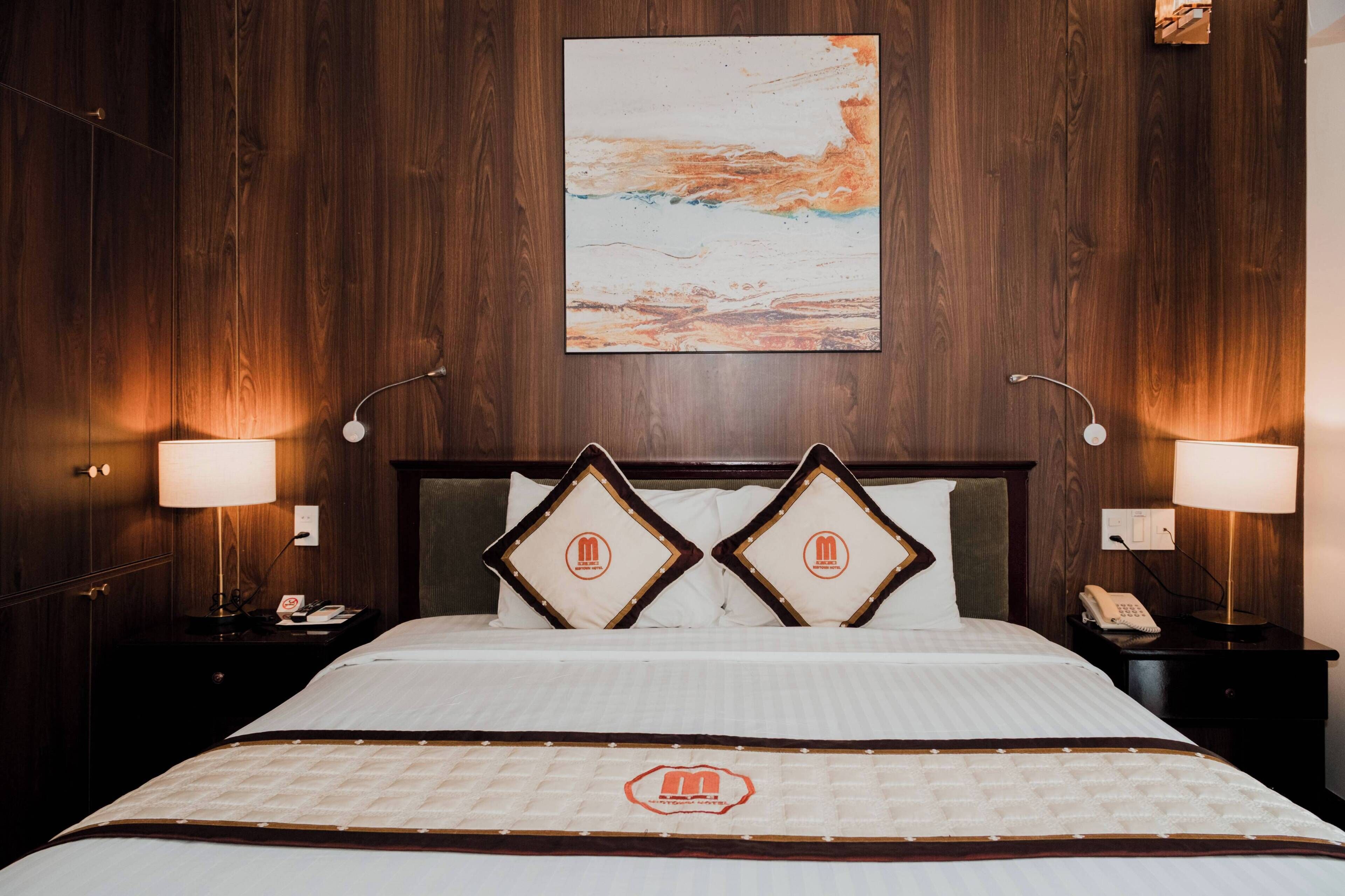 A hotel room with a wooden paneled wall, a large abstract painting, a bed with a quilted bedspread, two bedside lamps, and a telephone on the nightstand.