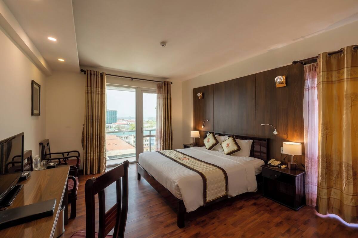 A hotel room with a large bed, a desk, and a chair. There is a window with curtains, a nightstand with a lamp, and a view of buildings outside.