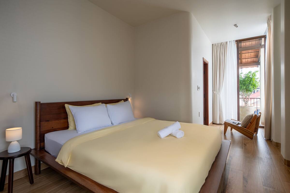 A neatly made bed with a wooden headboard, a bedside table with a lamp, and a chair by the window.