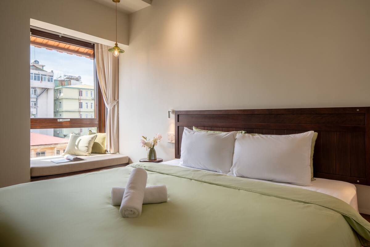 A hotel room with a large bed, a window with a view of buildings, a bedside table with a vase of flowers, and a wooden headboard.