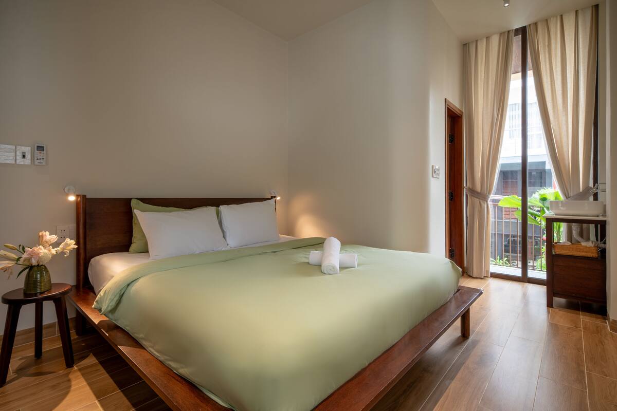 A hotel room with a bed, a bedside table with a vase, and a view of the outdoors.