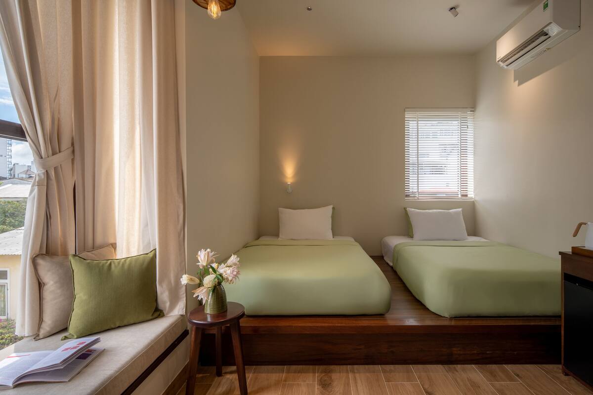 A hotel room with a bed, a bedside table, a window with blinds, and a view of the outside.