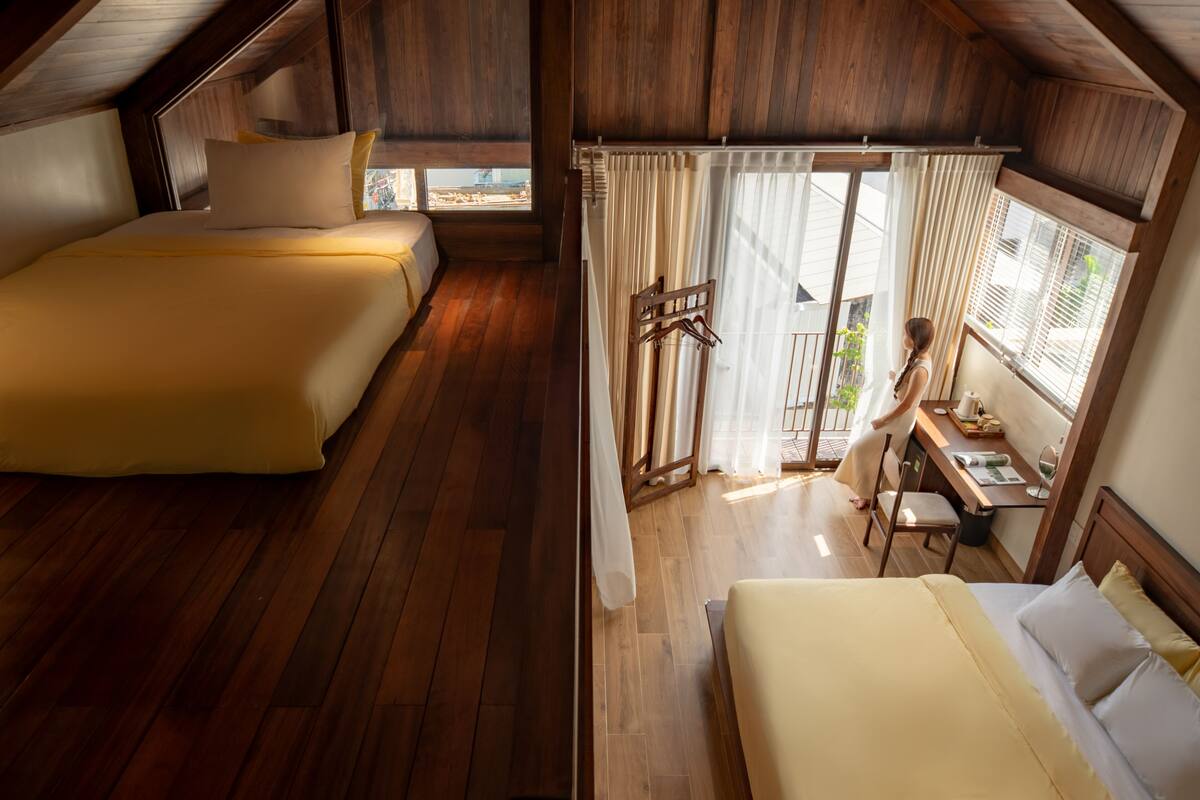 A bedroom with two beds, wooden floors, and a view of the outside through a window with curtains.