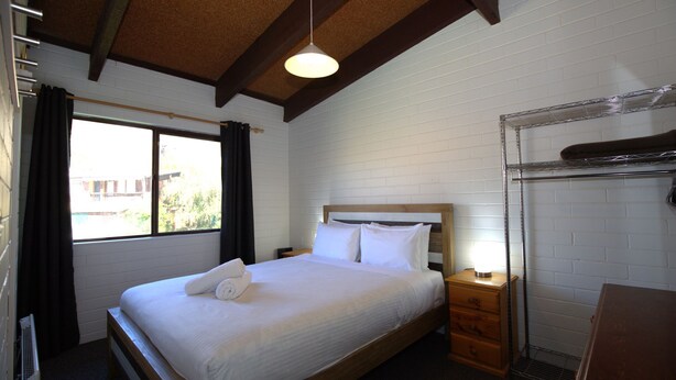 A hotel room with a large bed, wooden headboard, a nightstand, a dresser, a window with curtains, and a metal rack on the wall. - Alpha Centauri - 8/39 Gippsland Street (Jindabyne)