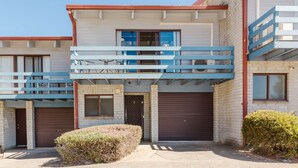 A modern two-story building with a balcony on the upper floor, a garage door on the ground floor, and a small bush in the front yard. - Alpha Centauri - 8/39 Gippsland Street (Jindabyne)