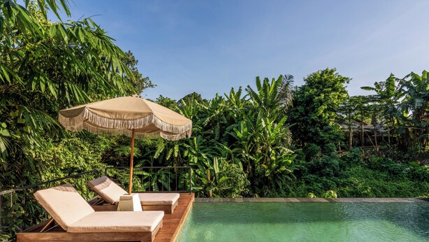 A wooden deck with two lounge chairs and a beige umbrella, overlooking a clear pool surrounded by lush greenery. - Jungle Breeze Villa by Betterplace (Cepaka)