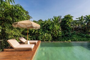Une terrasse en bois avec deux chaises longues et un parasol beige, donnant sur une piscine aux eaux cristallines, elle-même entourée d’une végétation luxuriante.